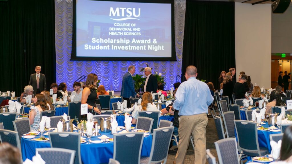 Ballroom setup for MTSU College of Behavioral and Health Sciences Scholarship Award & Student Investment Night, with blue tablecloths, formal dining setup, and stage with blue and gold draped backdrop and projector screen displaying event title.