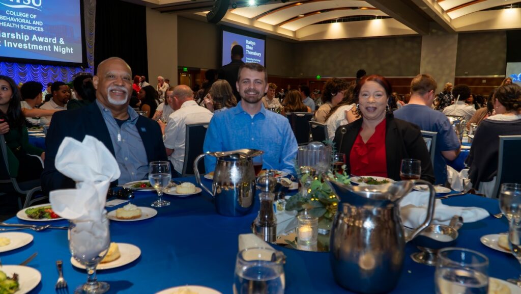 Three people smiling at dining table, with man in dark blazer on left, man in blue shirt in center, and woman in red blazer on right.