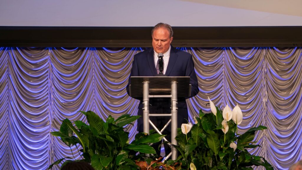 CBHS Dean, Peter Grandjean dark suit speaking at podium with microphone on stage with blue and gold draped backdrop.