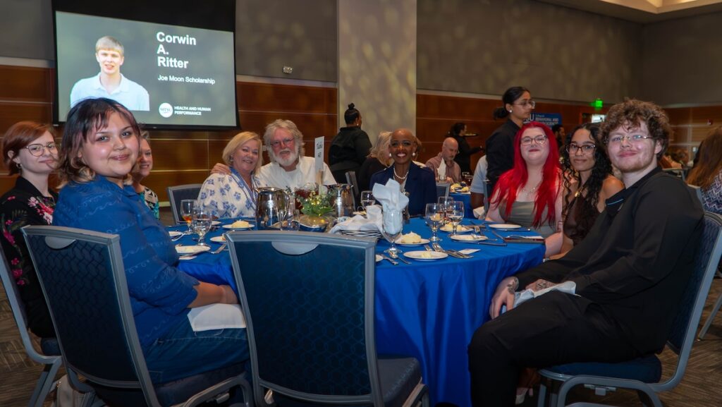people seated at blue table, with scholarship recipient name visible on screen above.