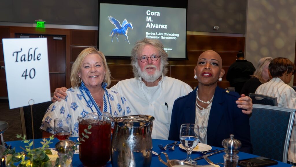 Three adults pose at a dining table during CBHS Student Awards Night. A presentation screen behind them displays "Cora M. Alvarez" and "Bertha & Jim Christzberg Recreation Scholarship." Table 40 is visible in the foreground.