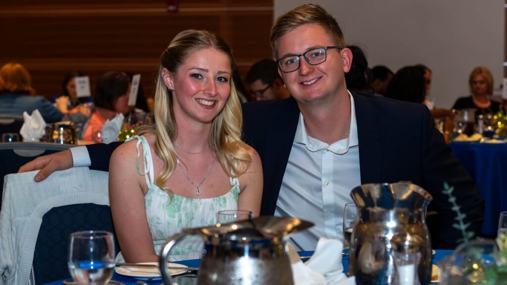 A young man and woman sit together at a blue-clothed dining table, both smiling at the camera during the awards dinner.