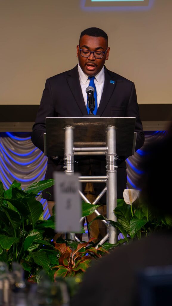RJ Ware speaks at a podium with a microphone. The stage is decorated with blue and tan draped fabric and green plants.