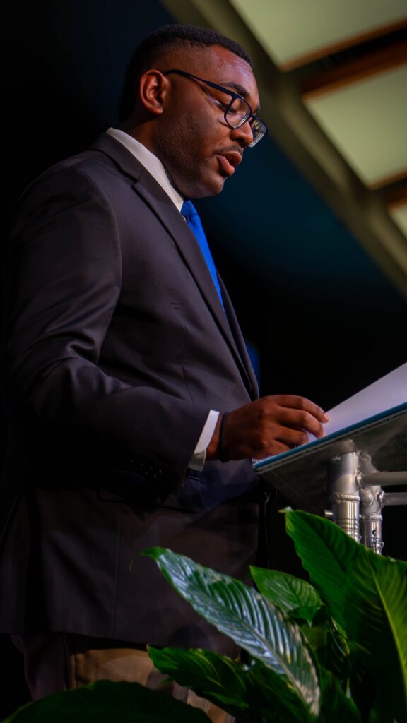 RJ Ware in a dark suit and blue tie at the podium during the ceremony.