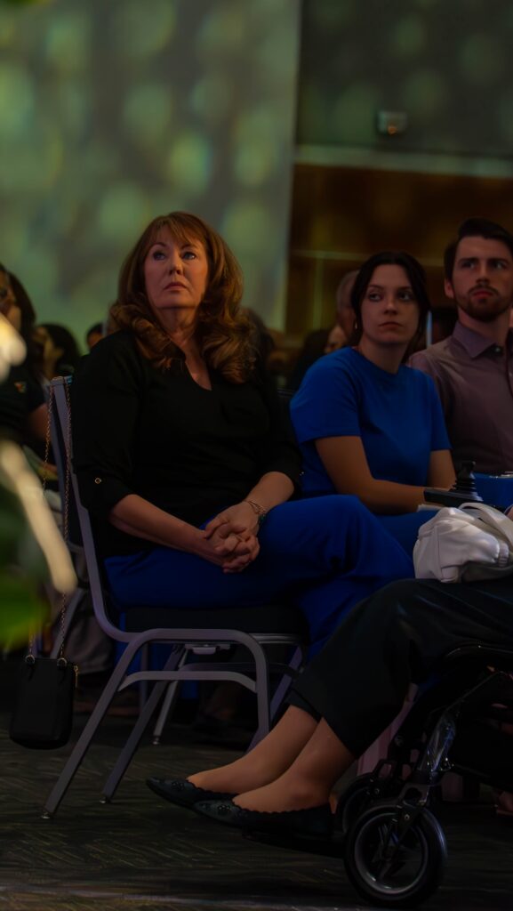 Three audience members sit in chairs watching the stage during the ceremony presentation.