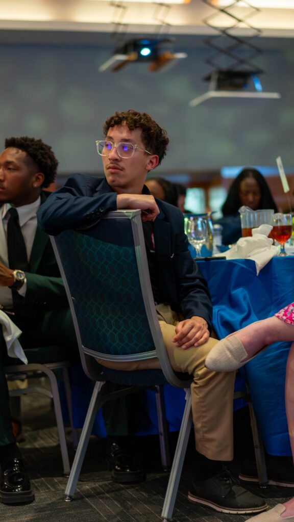 A young man in glasses and dark blazer sits at a dining table during the awards event.