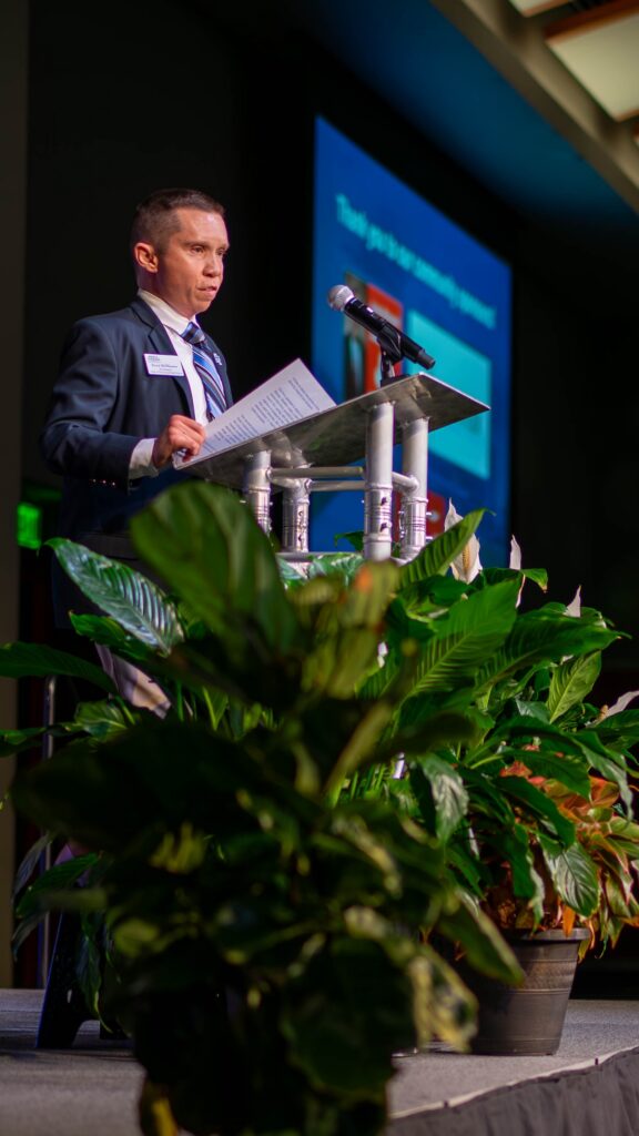 A man in a dark suit with blue tie holds papers at the podium during the ceremony.