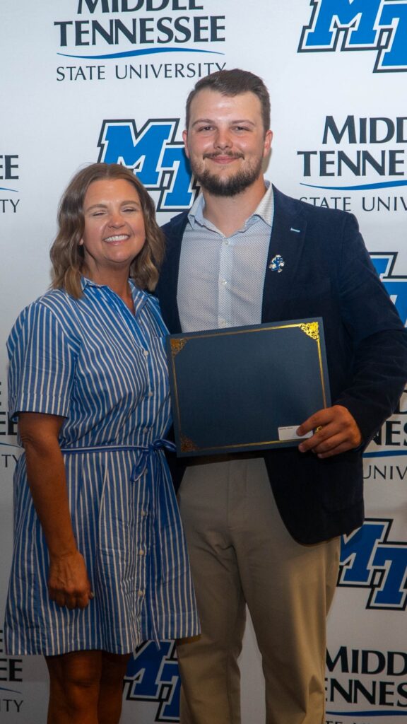 A woman in a blue striped dress and a man in a light blue button-up shirt and blazer stand in front of an MTSU backdrop, holding a framed scholarship certificate.