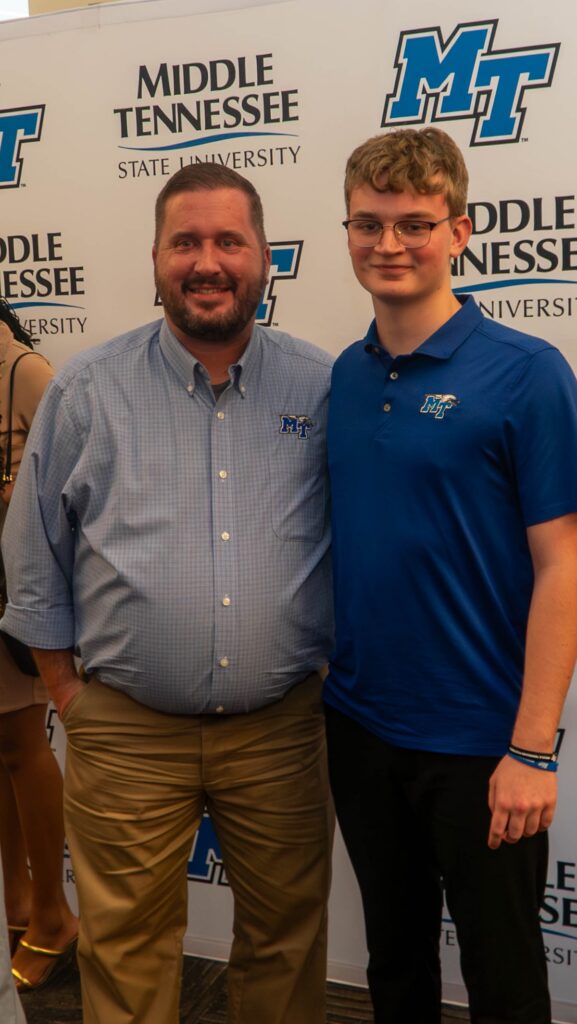 Two men stand together in front of an MTSU backdrop, both wearing MTSU branded apparel.