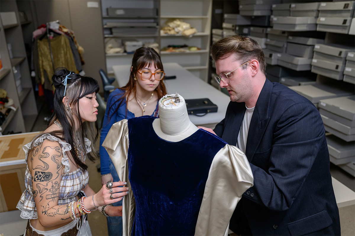 Three MTSU Fashion Studies students and faculty are gathered around a dress form in a design studio, closely examining a vintage-style blue velvet dress with cream-colored satin sleeves. On the left, a woman with dark hair and arm tattoos leans in to look at the fabric. In the center, a woman with reddish-brown hair and orange-rimmed glasses observes the process. On the right, a man in a dark blazer and glasses adjusts the garment on the mannequin. The background is filled with rows of organized grey storage boxes and shelving units.