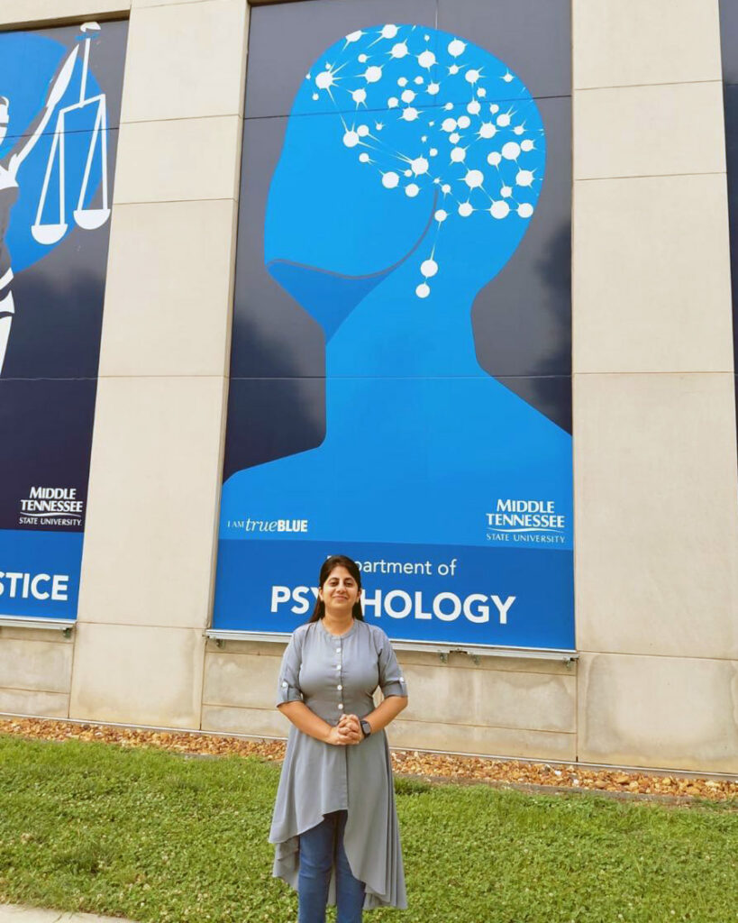 Dr. Meenakshi Shukla standing in front of the Psychology Department banner of the Academic Classroom Building at MTSU.
